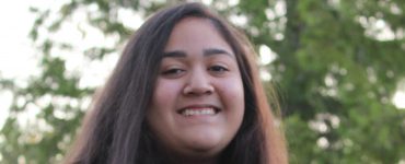 The face and upper body of a teenage girl, with long, straight, brown hair, brown eyes, and dark skin, wearing a sports sweatshirt and she is smiling.