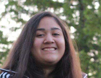 The face and upper body of a teenage girl, with long, straight, brown hair, brown eyes, and dark skin, wearing a sports sweatshirt and she is smiling.