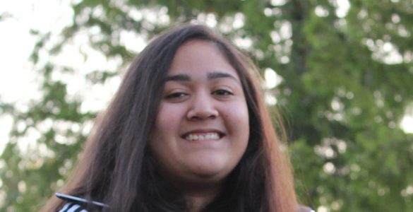 The face and upper body of a teenage girl, with long, straight, brown hair, brown eyes, and dark skin, wearing a sports sweatshirt and she is smiling.