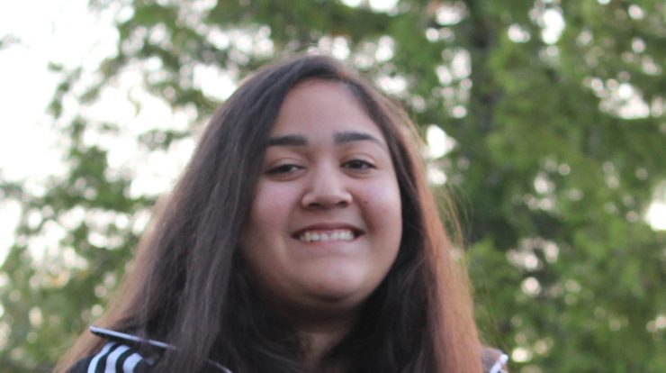 The face and upper body of a teenage girl, with long, straight, brown hair, brown eyes, and dark skin, wearing a sports sweatshirt and she is smiling.