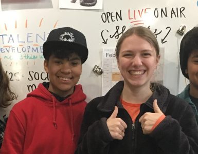 Four students standing in front of a large classroom white board. Two students are dark-skinned and appear to be identical twins. The other two students have lighter skin. Two students have wide smiles and are giving the thumbs up sign. Words on the white board behind the students include: daily schedule, talent development, and live on-air.