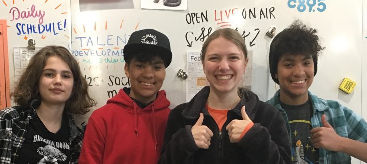 Four students standing in front of a large classroom white board. Two students are dark-skinned and appear to be identical twins. The other two students have lighter skin. Two students have wide smiles and are giving the thumbs up sign. Words on the white board behind the students include: daily schedule, talent development, and live on-air.