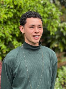 Photo of Ezra Conklin wearing a green turtle neck in front of green foliage