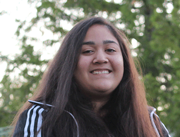 The face and upper body of a teenage girl, with long, straight, brown hair, brown eyes, and dark skin, wearing a sports sweatshirt and she is smiling.