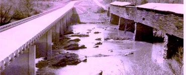 Black and white photo of a newly constructed bridge paralleling and older covered wooden bridge spanning a river.