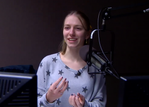 A photo of a young woman with medium length blond hair standing by a microphone. Her hands are in motion and she appears in mid-sentence.