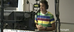 A student with dark skin and curly hair and a rainbow stripped shirt stands in front of a microphone emblazoned with the c895 logo