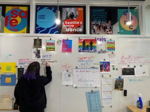 A photo of Harmony Soleil, writing on the whiteboard. The whiteboard is covered with motivational posters and information for students.