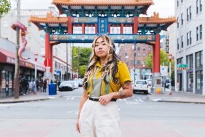 Artist Monyee Chau standing in the middle of the frame in front of the Historic Chinatown Gate in Seattle
