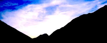 A blue sky with white clouds. The shadowy side of two mountains is in the foreground.