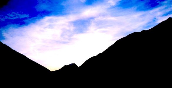 A blue sky with white clouds. The shadowy side of two mountains is in the foreground.