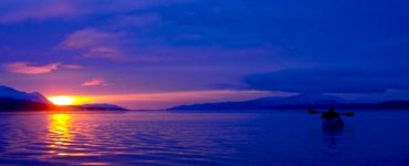 A person kayaking on a lake at sunset, with mountains in the background. The sky is a dark blue.