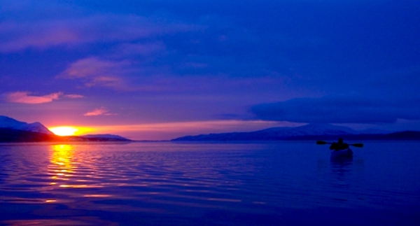 A person kayaking on a lake at sunset, with mountains in the background. The sky is a dark blue.