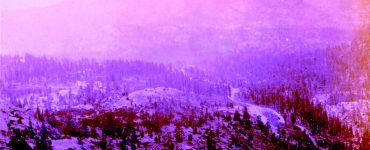 Old photograph of a hilly landscape with evergreen trees and snow. There is a valley running between the hills, and foggy weather conditions.