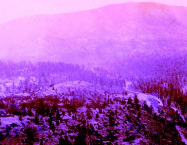 Old photograph of a hilly landscape with evergreen trees and snow. There is a valley running between the hills, and foggy weather conditions.