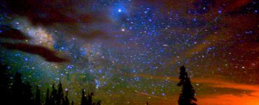 An image of a night sky in a rural area. The sky is star filled, with some clouds, and hues of blue and orange. There are shadows of trees in the foreground as well as evidence of human habitation in the form of a utility pole.