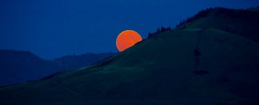 A deep orange moon rising from the mountain obscured horizon. The sky is dark blue. The mountains appear barren of vegetation, except for some evergreen trees near the peak.