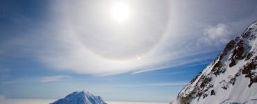 A halo around the sun, over a snowy landscape, with mountain peaks in the foreground. The sky is party cloudy but bright.