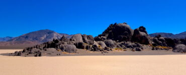 In the foreground is a flat dry lake bed, with a outcropping of rocks that somewhat resemble a grandstand at a race track. The sky is blue and there are mountains further in the desert background.