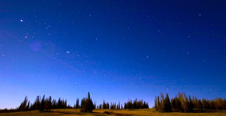 View of the night sky at sunset. The sky is becoming dark blue, and the sun is still illuminating the greenery on the ground. The sky is filled with stars, and gets deeper blue as the altitude increases.