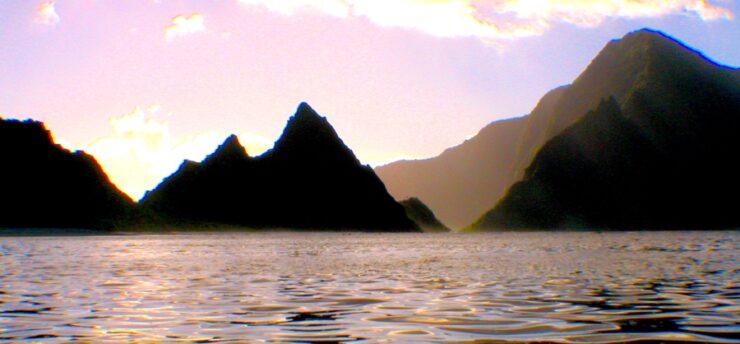 A body of water at dusk or dawn, with mountains jetting out of the water, forming islands or peninsulas. The sky is light purple and there is a light mist.