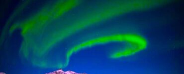 Florescent green aurora bands in a dark blue sky above a melting snowy polar landscape.