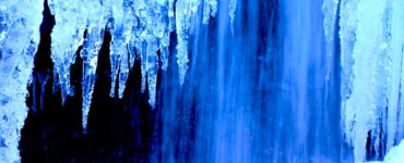 Icicles in the foreground with a waterfall in the background. The ice and water have a blue tint to them.