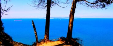 And sand-covered cliff edge with deciduous trees. The cliff is overlooking a large lake that is deep blue. The sky in the background is blue. There is some haze in the distance.