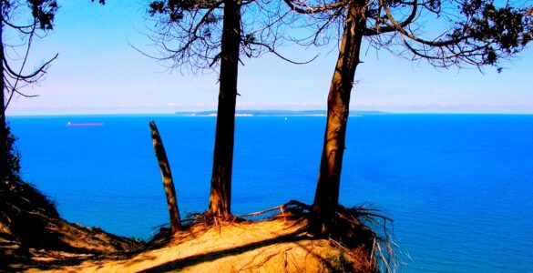 And sand-covered cliff edge with deciduous trees. The cliff is overlooking a large lake that is deep blue. The sky in the background is blue. There is some haze in the distance.