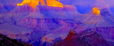 View of a canyon extending to the horizon. The canyon is shaded with sunlight reaching the peaks of the mountains within the canyon. In the foreground is the shaded edge of a cliff. The sky is blue with some white clouds on the horizon.