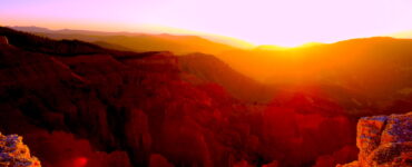 The sun is setting behind a ridge of mountains. In the foreground is the edge of a cliff exposing the jagged, but beautiful landscape below. Another range of mountains falls between the cliff and the ridge with the sunset. The landscape is orange, and the sky is shades of green and purple, with a yellow and orange sunset.