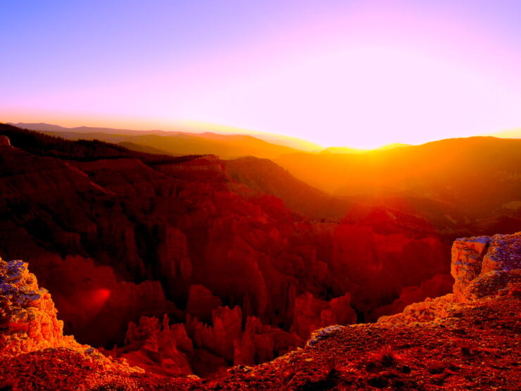 The sun is setting behind a ridge of mountains. In the foreground is the edge of a cliff exposing the jagged, but beautiful landscape below. Another range of mountains falls between the cliff and the ridge with the sunset. The landscape is orange, and the sky is shades of green and purple, with a yellow and orange sunset.