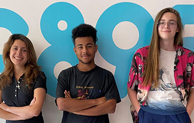 A photo of three teenagers standing in front of a white wall with the C89.5 logo. The person on the left has long brown hair and tan skin and has their arms folded. The person in the middle has dark skin and black curly hair and their arms crossed. The person on the right has long brown hair, glasses, pale skin and has their hands in their pockets. They all looked relaxed and are smiling.