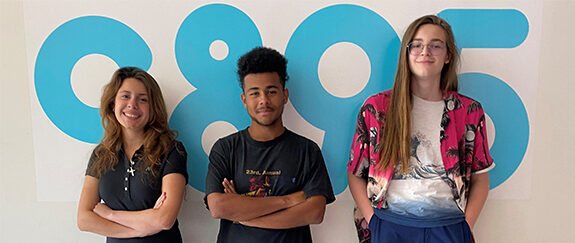 A photo of three teenagers standing in front of a white wall with the C89.5 logo. The person on the left has long brown hair and tan skin and has their arms folded. The person in the middle has dark skin and black curly hair and their arms crossed. The person on the right has long brown hair, glasses, pale skin and has their hands in their pockets. They all looked relaxed and are smiling.