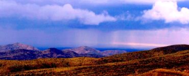 A desert landscape featuring a distance rainstorm. In the foreground are short, orange hills without vegetation. Behind that are taller hills that appear blue, and are also without vegetation. Behind that is a blue cloudy sky, with two curtains of rain reaching the landscape.