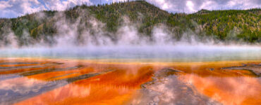 A volcanically active body of water with steam rising above. The water in the foreground appears orange or rust colored. The water further away appears to be blue. In the background are hills with green and brown foliage. The sky conditions are mostly cloudy, with sections of blue peeking through.