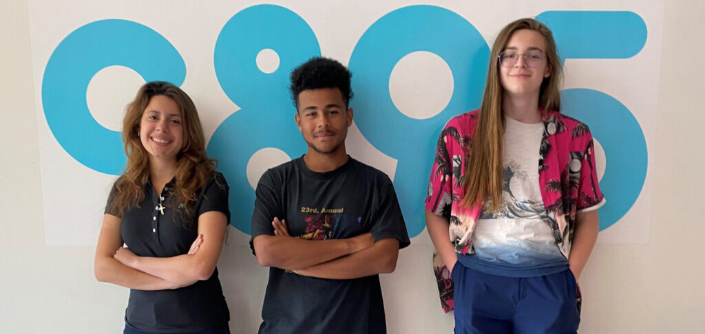 A photo of three young people standing in front of the c89.5 logo on a wall. One of the students has tan skin, long brown hair and is smiling folding her arms casually, Another student has dark skin, curly black hair and is smiling. The third student is tall, has pale skin, long hair and is wearing glasses. They have their hands in their pockets and is smiling.