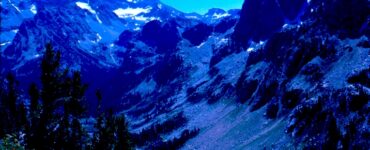 A canyon with minimal vegetation and tiny bits of snow near the valley mountain peaks. In the foreground are wild flowers and a few evergreen trees. The sky is blue.