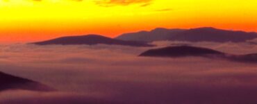 A pre-sunrise landscape, with mountaintops in view. There is a valley below that you can not see, as it is covered with fog, so it appears that the photo is taken above the clouds. The mountain tops are dark, and the background sky is illuminated by the forthcoming sun, with yellow and orange hues.