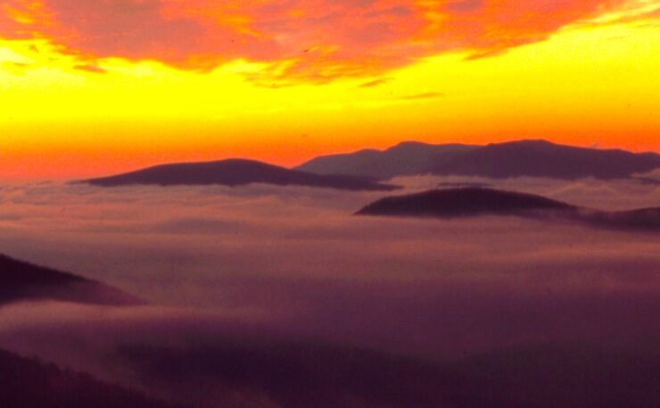 A pre-sunrise landscape, with mountaintops in view. There is a valley below that you can not see, as it is covered with fog, so it appears that the photo is taken above the clouds. The mountain tops are dark, and the background sky is illuminated by the forthcoming sun, with yellow and orange hues.