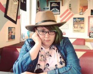 An image of Megan Garbayo-López sitting at a diner, wearing a tan wide brimmed hat wearing glasses with hair that is dyed green.