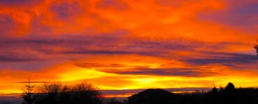 A bright sunset is illuminated on a mostly cloudy sky, with hues of orange and yellow. Parts of the clouds are dark blue. In the foreground, on the lower-third, is a building and some deciduous trees with no leaves, and a sole evergreen tree. There is also a cabin the the foreground. The foreground is shown as mostly an outline, as the sun is not providing light on that area.