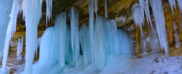 Icicles hanging from below an escarpment. The icycles are robust and light blue. There is white ice on the ground.