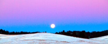 In the foreground is a landscape with flowing hills and no vegetation. The hills are covered in a shallow blanket of snow. Behind the hills are a line of trees, about a quarter mile away. The sky shows a bright moon about to set on the horizon. The sky is blue and purple.