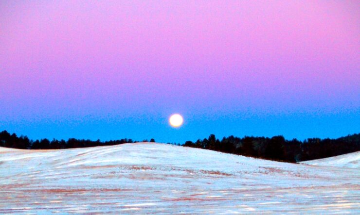In the foreground is a landscape with flowing hills and no vegetation. The hills are covered in a shallow blanket of snow. Behind the hills are a line of trees, about a quarter mile away. The sky shows a bright moon about to set on the horizon. The sky is blue and purple.