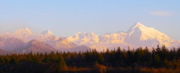 In the foreground is a lake with calm blue waters. The shore of the lake is forested, mostly with evergreen trees. In the background are tall mountains, the ones the furthest away are snow covered. The sky is blue with a three-quarter moon showing in the daylight.