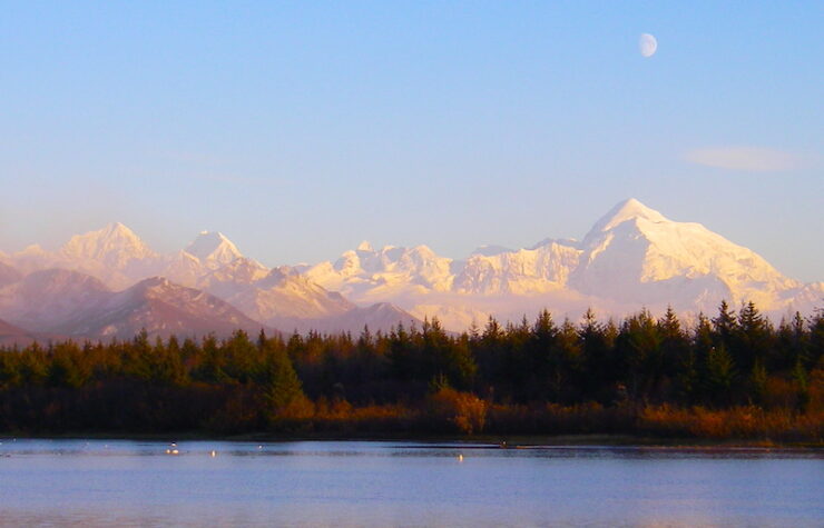 In the foreground is a lake with calm blue waters. The shore of the lake is forested, mostly with evergreen trees. In the background are tall mountains, the ones the furthest away are snow covered. The sky is blue with a three-quarter moon showing in the daylight.