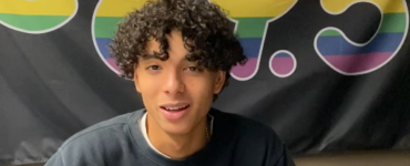 A photo of a teenage boy. He has light brown skin, brown curly hair and is smiling.