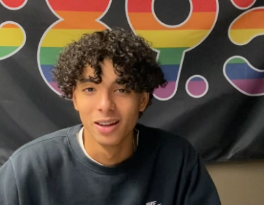 A photo of a teenage boy. He has light brown skin, brown curly hair and is smiling.