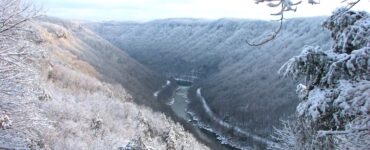 Lightly snow-covered landscape of a gorge with a winding river and steep hillsides. Snow on tree branches in foreground. Mostly cloudy sky.